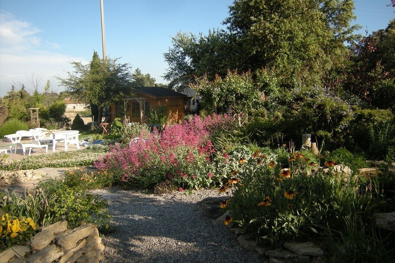 Un jardin avec un chemin de gravier, différentes plantes et un bâtiment en arrière-plan.