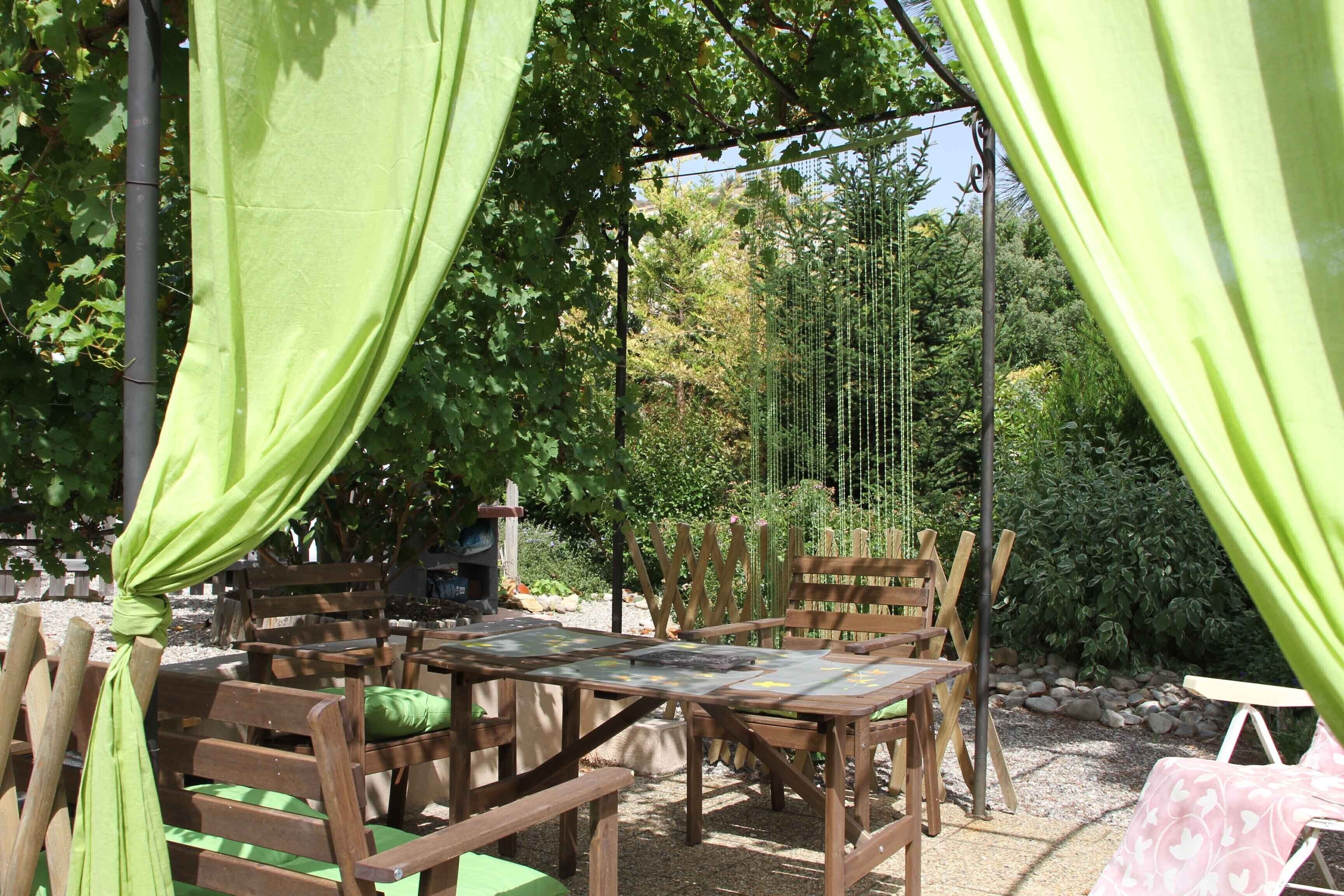 Un espace extérieur aménagé avec des meubles en bois et des coussins verts, entouré de verdure et surmonté d’une pergola.