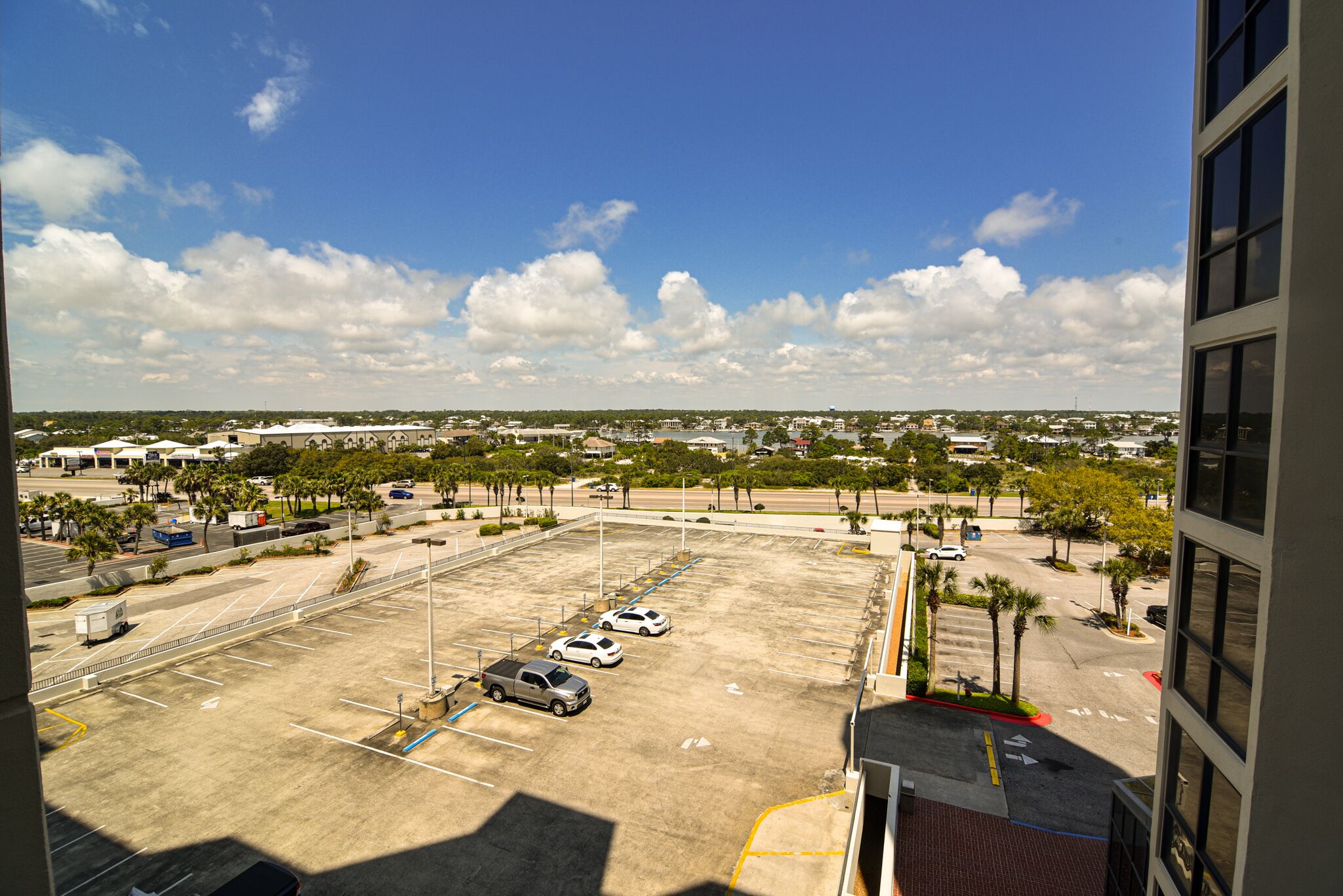 Un parking avec plusieurs voitures, un bâtiment doté de grandes fenêtres et un ciel dégagé avec quelques nuages.