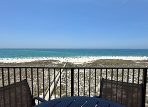 A beach view from a balcony with a table and chairs, a sandy beach, and the ocean.