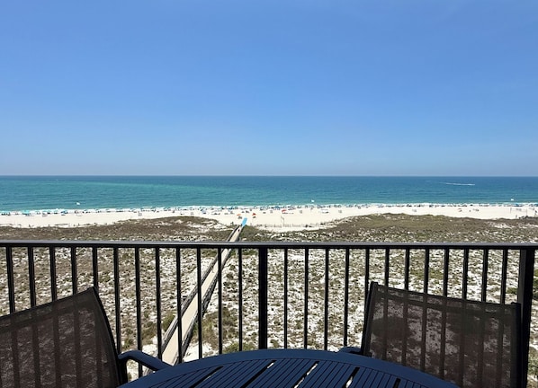 A beach view from a balcony with a table and chairs, a sandy beach, and the ocean.
