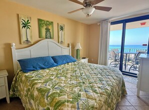 Habitación de hotel con cama, ventilador de techo, mesita de noche, lámpara y vista al océano desde un balcón.