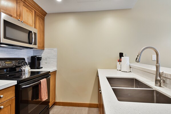 A modern kitchen with wooden cabinets, a stainless steel microwave, a kettle, and a sink with a faucet.