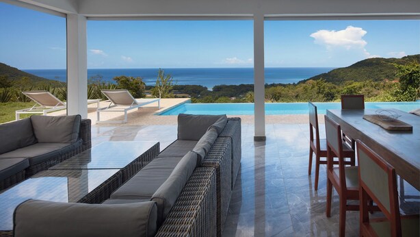 Un espace extérieur moderne, aménagé avec une table et des chaises en verre, offrant une vue sur une piscine et l’océan.