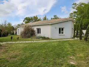 Uma casa térrea com fachada cinza clara, telhado ladrilhado e um jardim bem cuidado com arbustos e árvores.