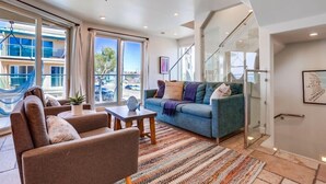 Ein modernes Wohnzimmer mit Sofa, Sesseln und Couchtisch. Ein großes Fenster mit Vorhängen, ein Deckenventilator und Blick auf Gebäude.