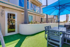 An outdoor patio with a table and chairs, a blue umbrella, and a white wall with a window.