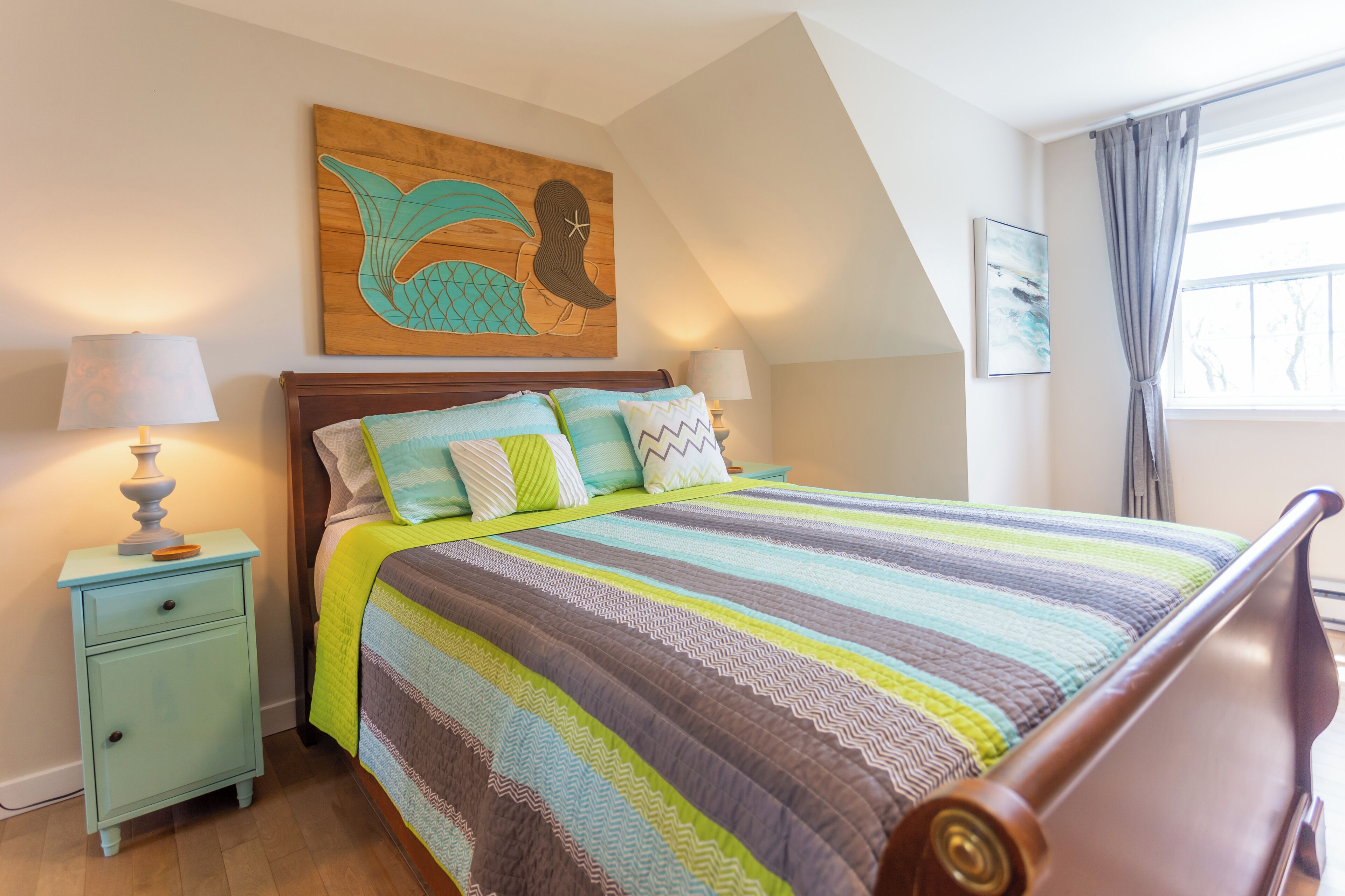 A bedroom with a wooden bed, striped bedding, a nightstand with a lamp, and a mermaid artwork on the wall.