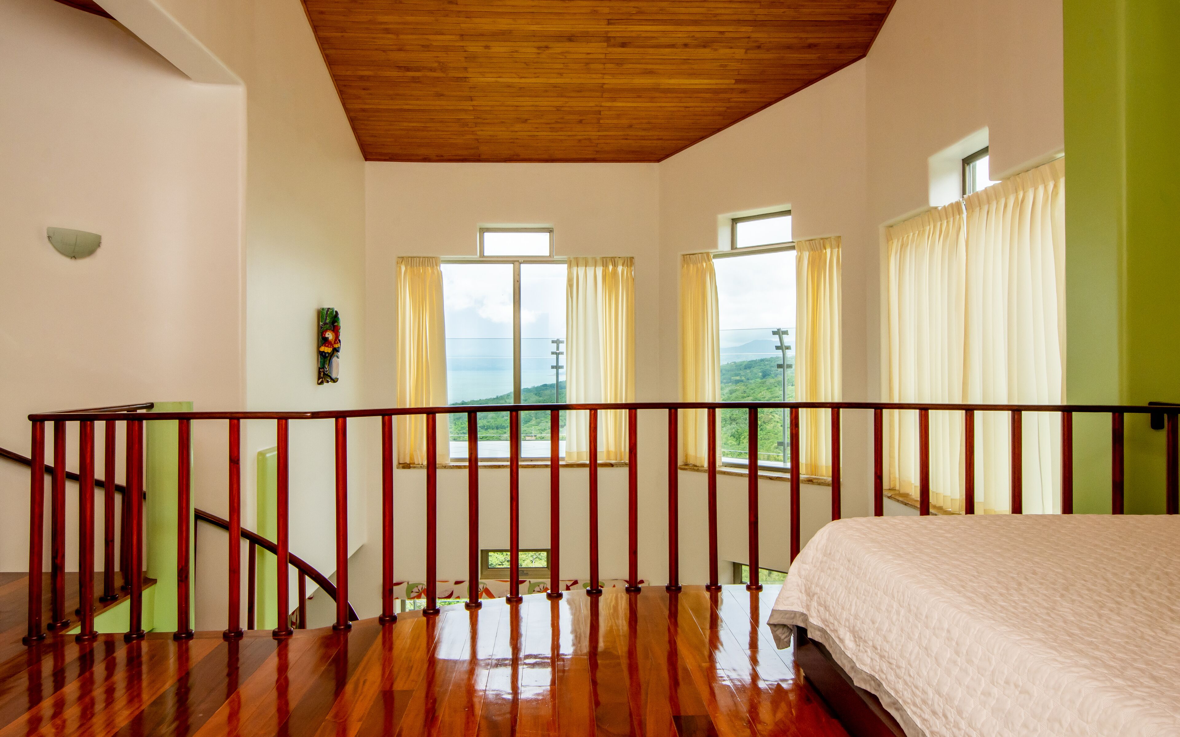 A bedroom with a wooden ceiling, a bed with a white duvet, a wooden staircase, and large windows with curtains.