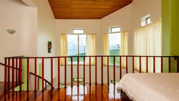 A bedroom with a wooden ceiling, a bed with a white duvet, a wooden staircase, and large windows with curtains.