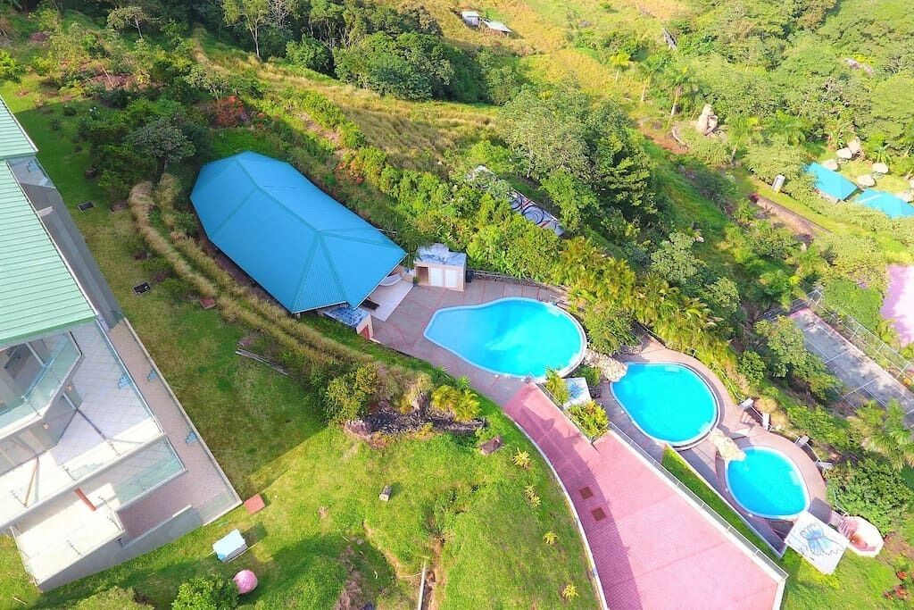 Vue aérienne d’une propriété dotée de plusieurs piscines, d’un grand espace couvert et d’une végétation luxuriante.