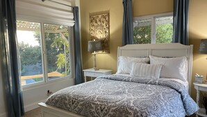 A bedroom with a large bed, two bedside tables, a lamp, and a view of greenery outside. - Views, Ocean/Mountain, Hot Tub, AC, Walk to Mesa Village, Gated Yard (Santa Barbara)