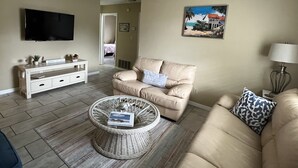 A living room with a beige sofa, a wicker coffee table, a white entertainment unit, a flat-screen TV, and a painting on the wall. - Get To the Beach!  Only 5 miles to Siesta Key Beach! (Sarasota)