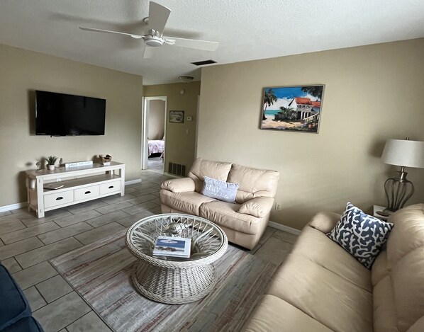 A living room with a beige sofa, a wicker coffee table, a white entertainment unit, a flat-screen TV, and a painting on the wall. - Get To the Beach!  Only 5 miles to Siesta Key Beach! (Sarasota)