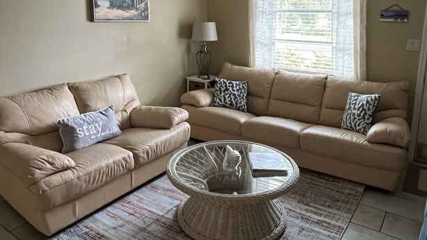 A living room with two beige sofas, a glass coffee table, a painting of a beach scene, a lamp, and a window with curtains. - Get To the Beach!  Only 5 miles to Siesta Key Beach! (Sarasota)