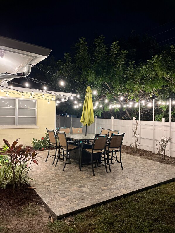 An outdoor patio with a table and chairs, surrounded by a white fence and string lights. - Get To the Beach!  Only 5 miles to Siesta Key Beach! (Sarasota)