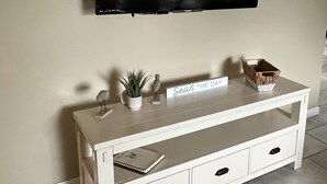 A white TV stand with a flat-screen TV mounted above it, a small potted plant, a decorative bird, a sign that reads "Seas THE DAY," and a basket. - Get To the Beach!  Only 5 miles to Siesta Key Beach! (Sarasota)