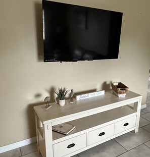 A white TV stand with a flat-screen TV mounted above it, a small potted plant, a decorative bird, a sign that reads "Seas THE DAY," and a basket. - Get To the Beach!  Only 5 miles to Siesta Key Beach! (Sarasota)