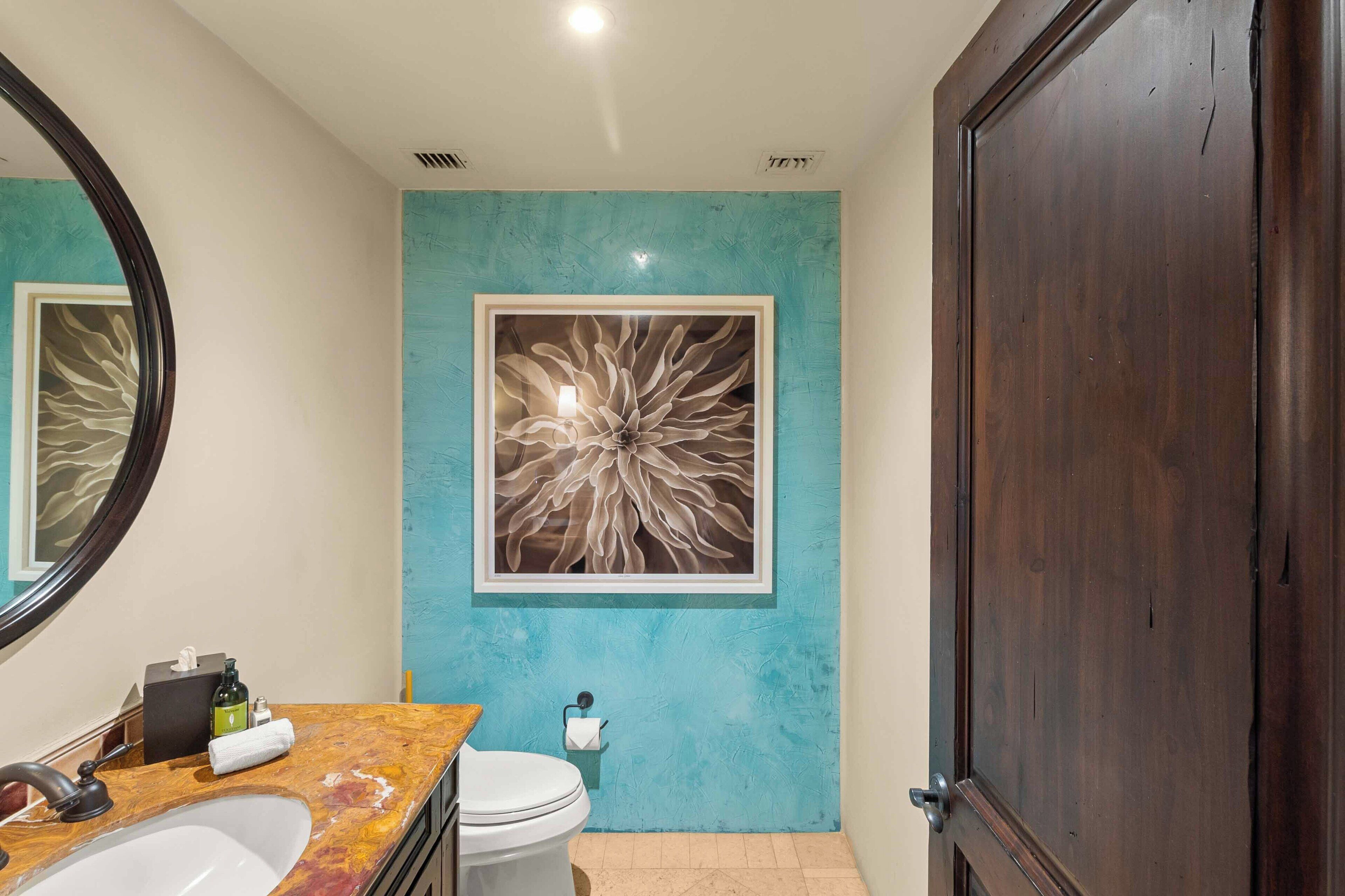 Un baño con lavamanos de mármol, inodoro, una obra de arte abstracto enmarcada en la pared y una puerta de madera oscura.