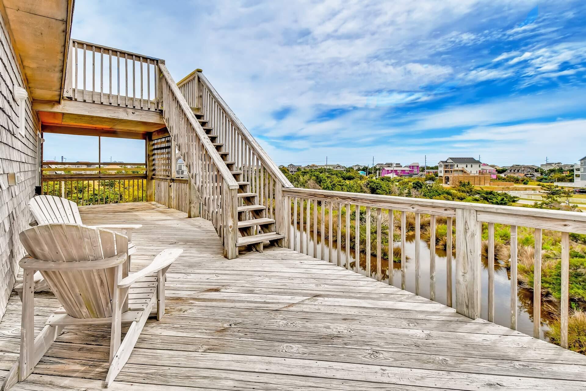 A wooden deck with a staircase leading to a covered area, overlooking a body of water and residential buildings.