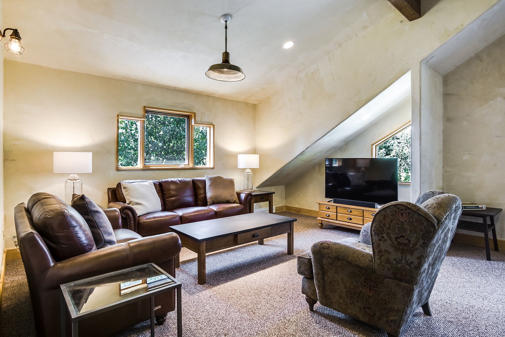 A spacious living room with a brown leather sofa, a patterned armchair, a glass coffee table, and a flat-screen TV. Large windows provide natural light and a view of greenery outside.