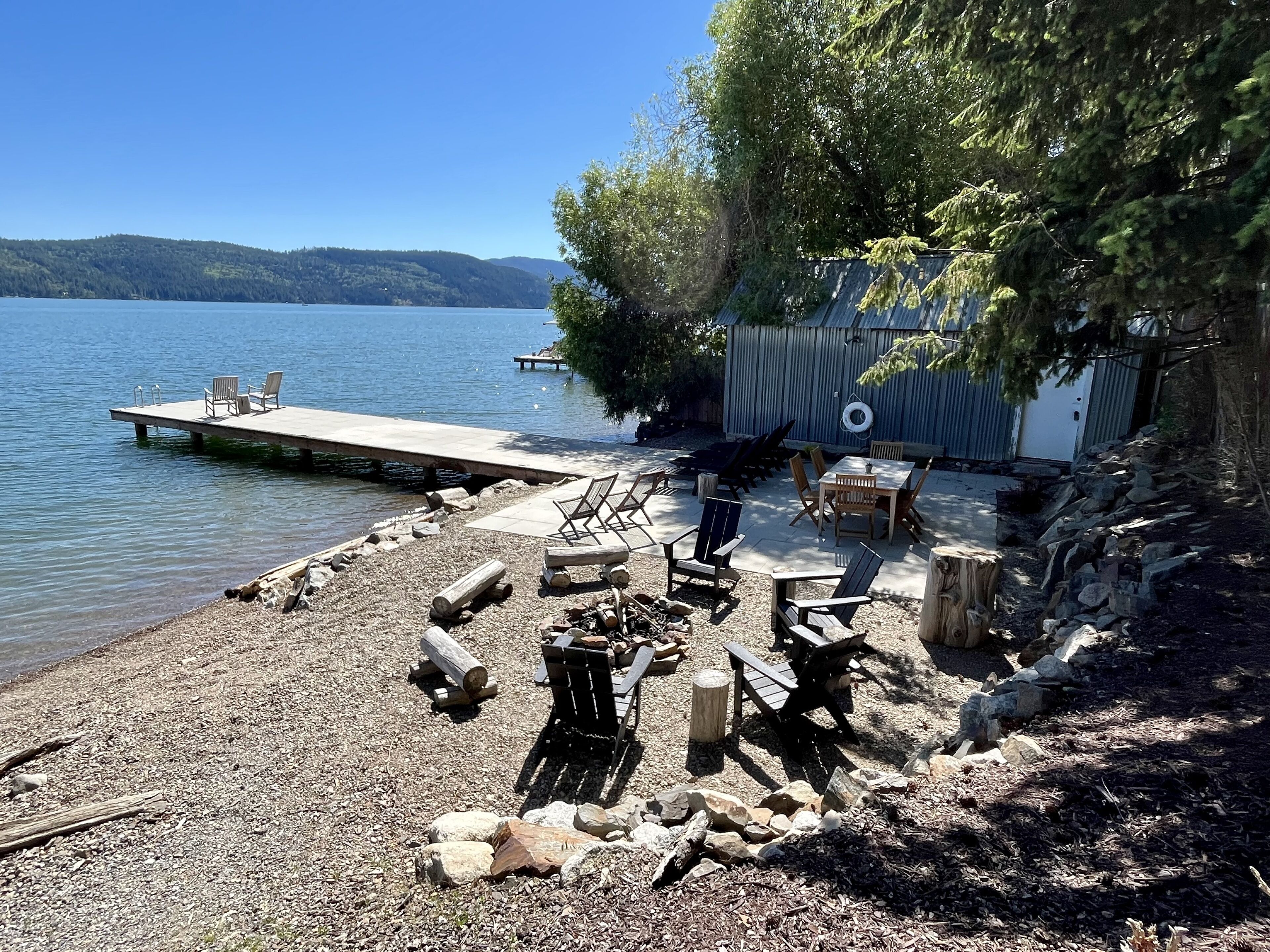 A lakeside cabin with a dock extending into the water, surrounded by a gravel beach and wooden furniture.