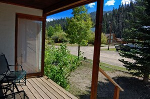 Une terrasse en bois avec une porte vitrée donnant sur une pièce, et offrant une vue sur une zone boisée avec un sentier de terre et un bâtiment situé au loin.