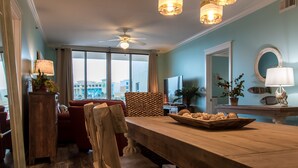 A spacious living room with a wooden dining table, red sofas, and a large window offering a city view. - 🦩Charming Walk-Out Level W/ Upper Pools~No Elevator Needed! Waterscape A233🦩 (Fort Walton Beach)