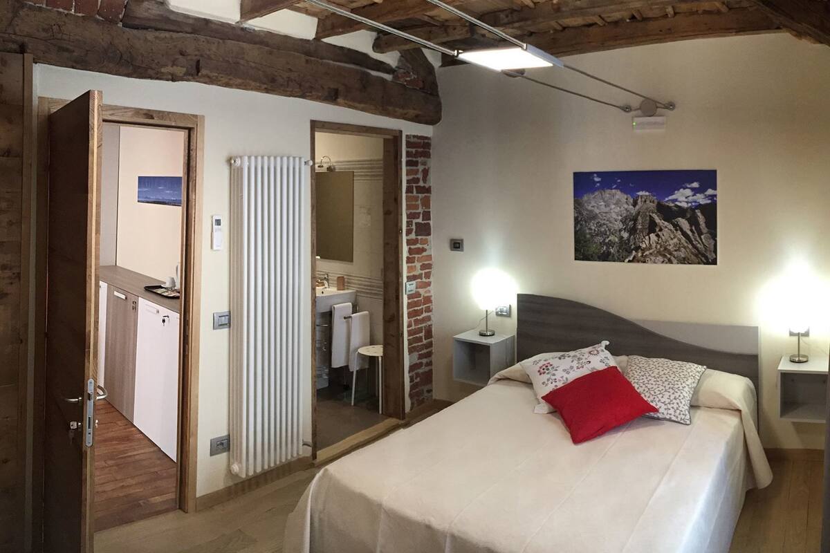A bedroom with a bed, a nightstand, a wall-mounted lamp, a radiator, a bathroom visible through an open door, and a painting on the wall.