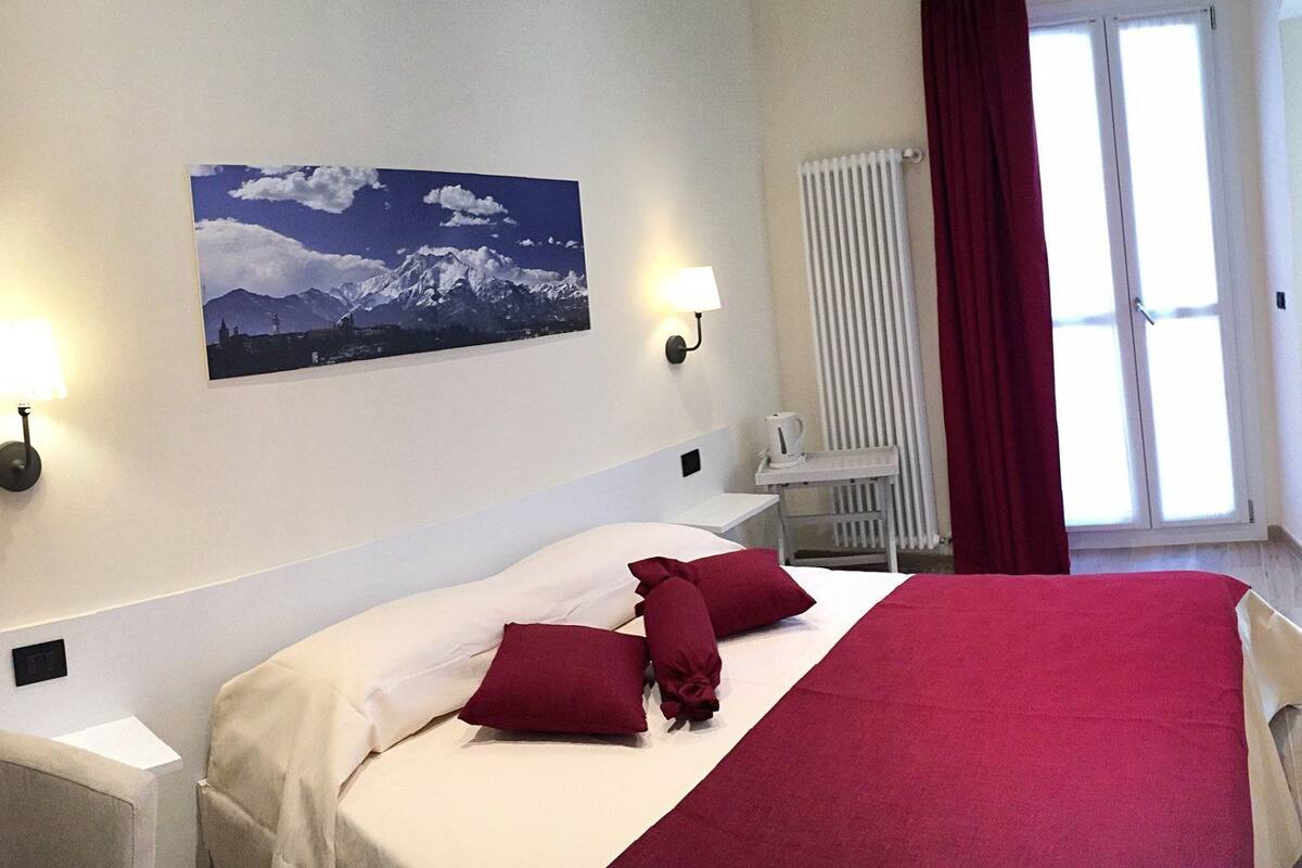 A hotel room with a bed, two bedside lamps, a radiator, a window with curtains, and a wall-mounted picture.
