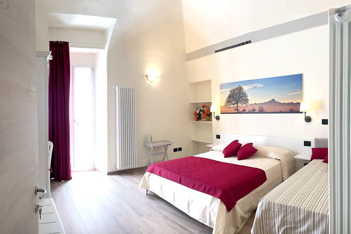 A hotel room with a large bed, a smaller bed, a radiator, a desk with a chair, a window with curtains, and a wall-mounted picture.