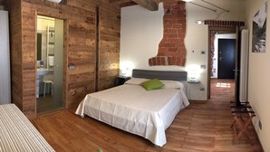 A bedroom with a bed, a wooden floor, a brick wall, and a bathroom visible through a glass door. - Osteria Senza Fretta Affittacamere (Cuneo)