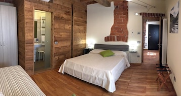 Un dormitorio con cama, suelo de madera, pared de ladrillo y baño visible a través de una puerta de vidrio.