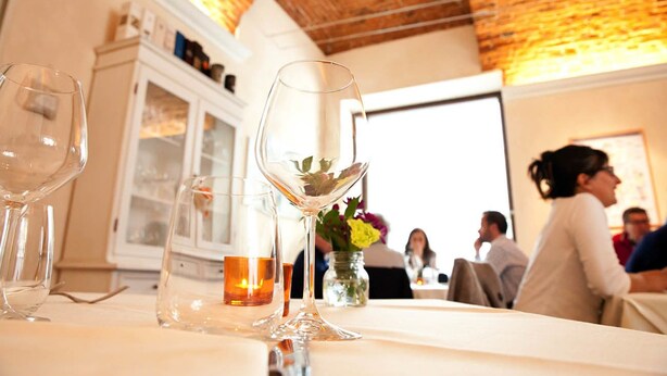 A table with wine glasses and a candle, set in a room with a brick ceiling and white cabinets in the background. - Osteria Senza Fretta Affittacamere (Cuneo)