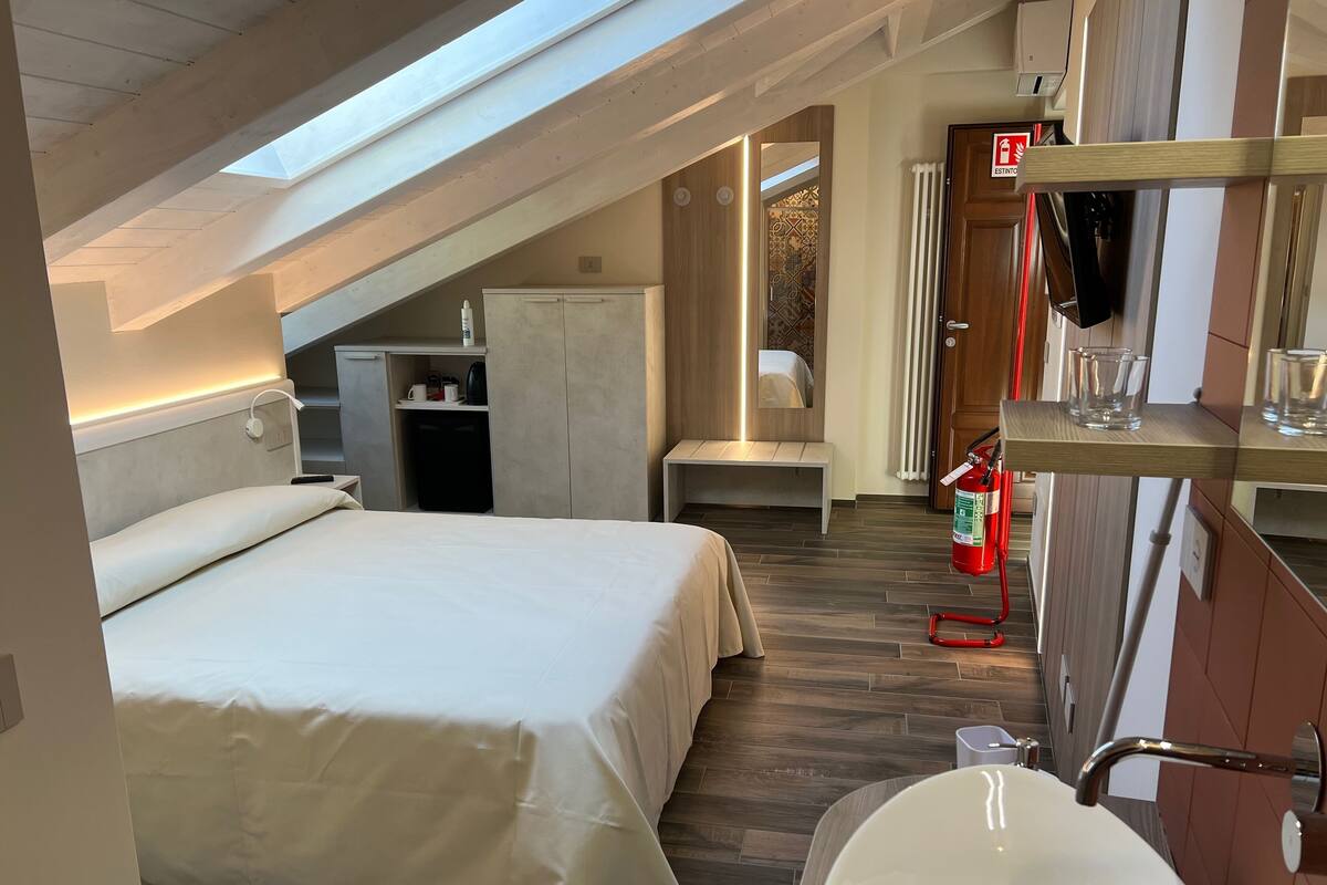 A bedroom with a skylight, a bed, a small desk, a bench, and a bathroom with a sink and mirror.