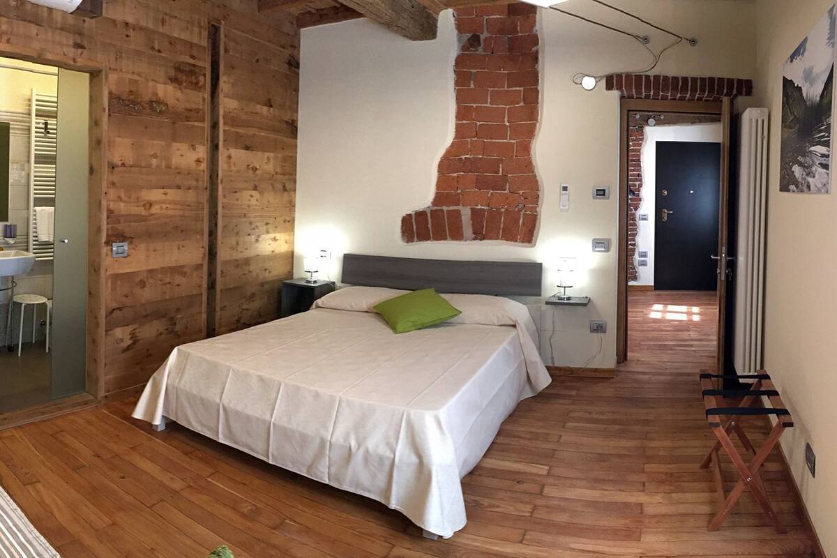 A bedroom with a wooden floor, a bed with a white bedspread, a green pillow, a nightstand with a lamp, and a bathroom visible through a glass door.
