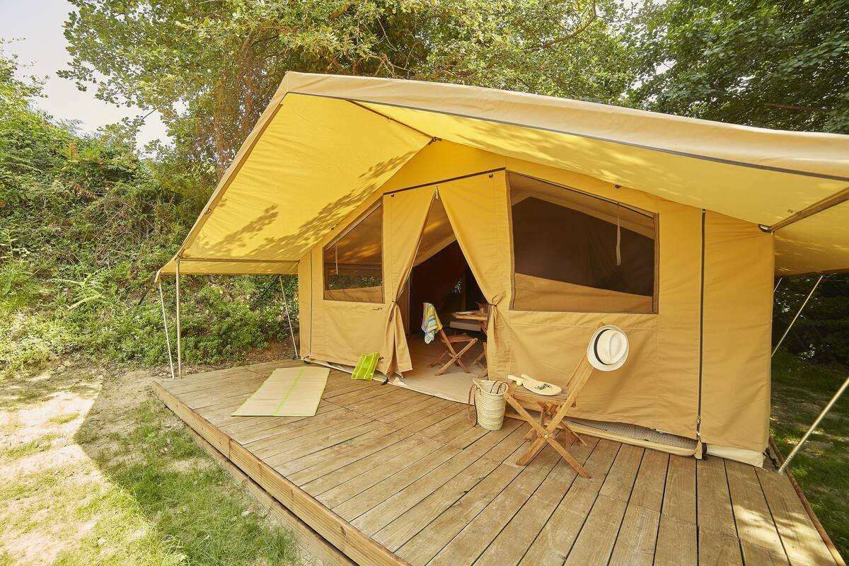 Una tienda amarilla con plataforma de madera, una silla plegable y un sombrero colgado al lado.