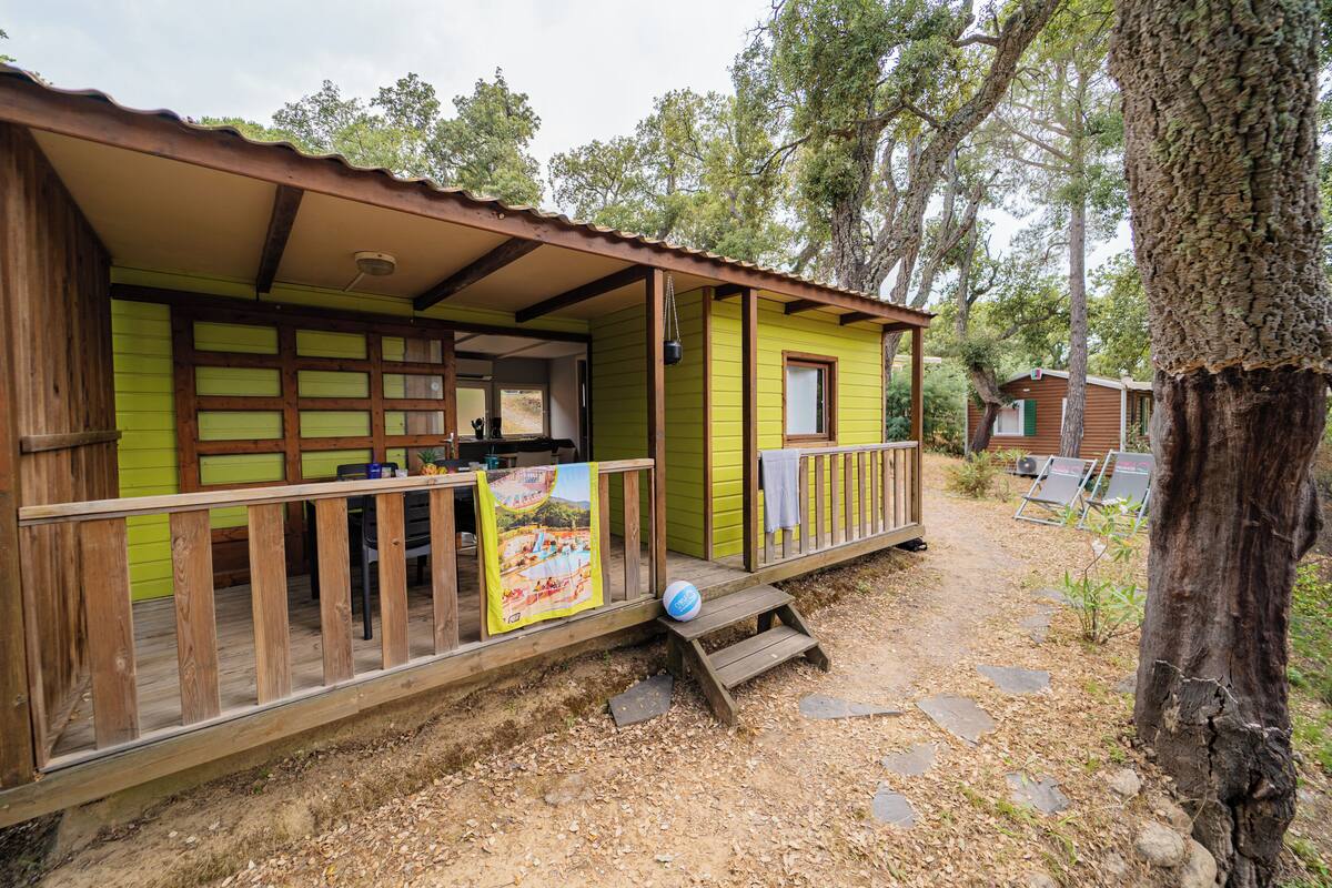 Una cabaña de madera con un porche, una pequeña terraza y un sendero de tierra que conduce a otra cabaña.