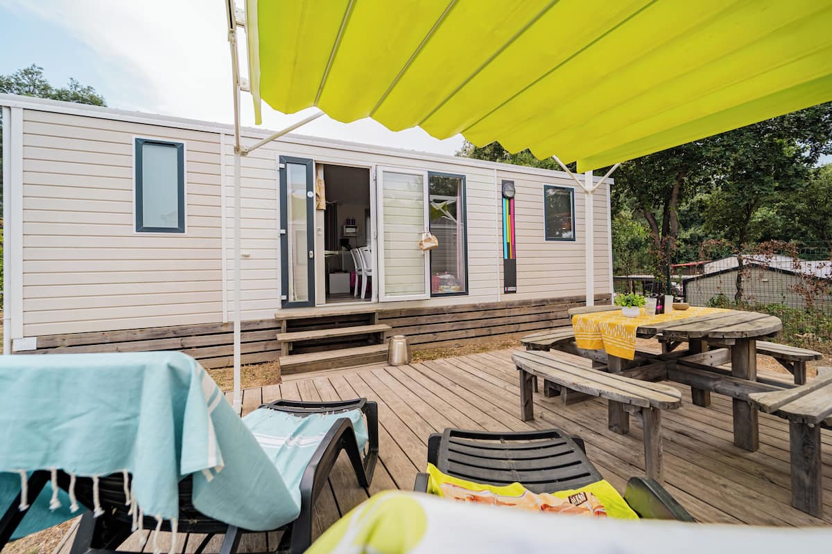Una casa móvil con terraza, mobiliario exterior y una mesa de picnic techada.