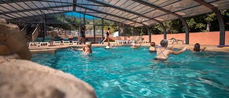 Une piscine dotée d’un toit en verre, entourée de chaises longues et d’un mur en pierre.