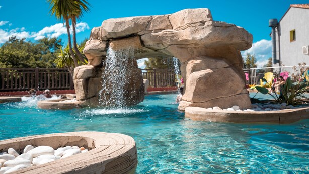 Une piscine agrémentée d’une arche en pierre et d’un élément décoratif aquatique, entourée de palmiers et d’une clôture.