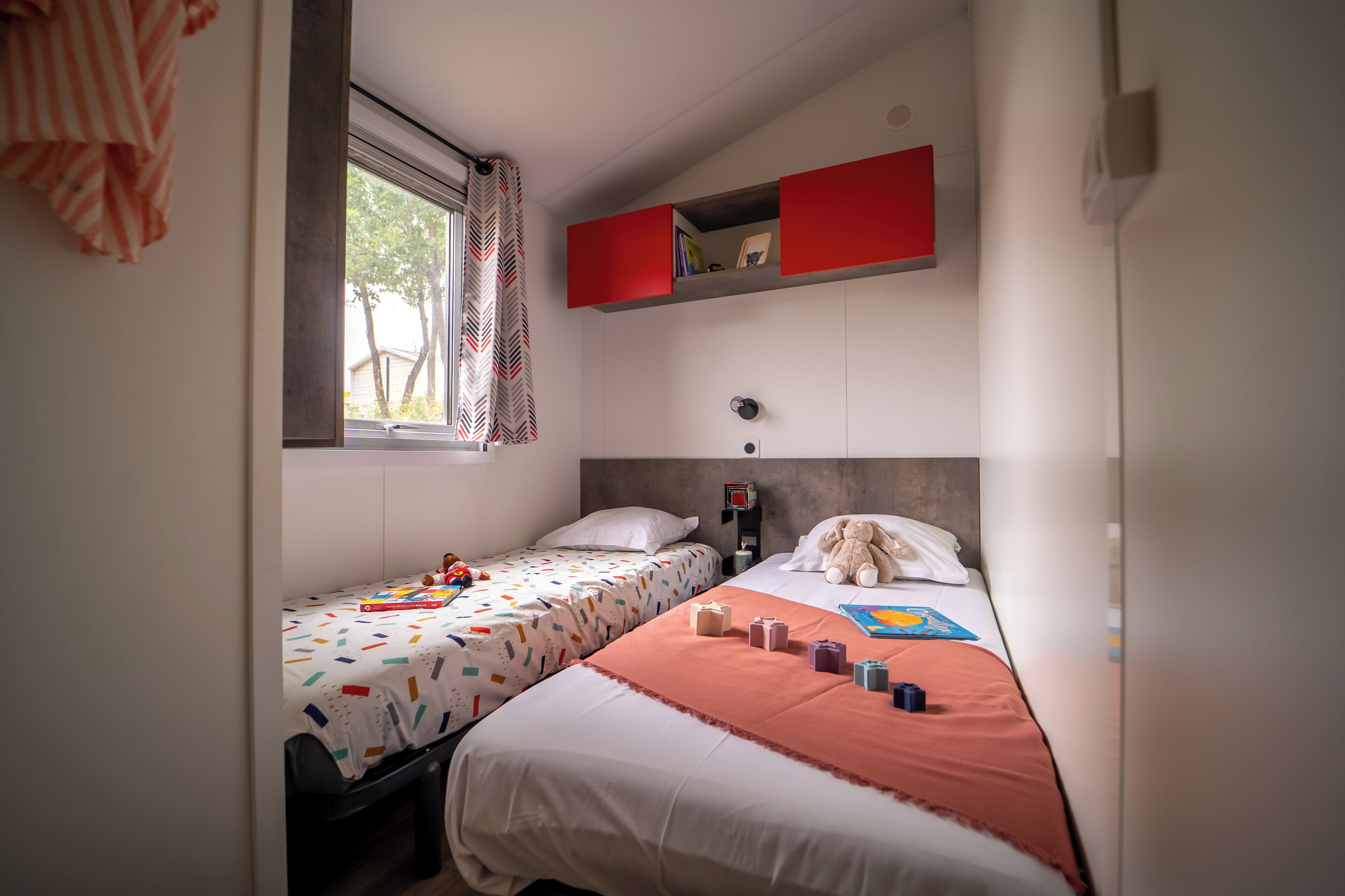 A bedroom with two beds, a window with curtains, a red shelf above the beds, and a teddy bear on the right bed.