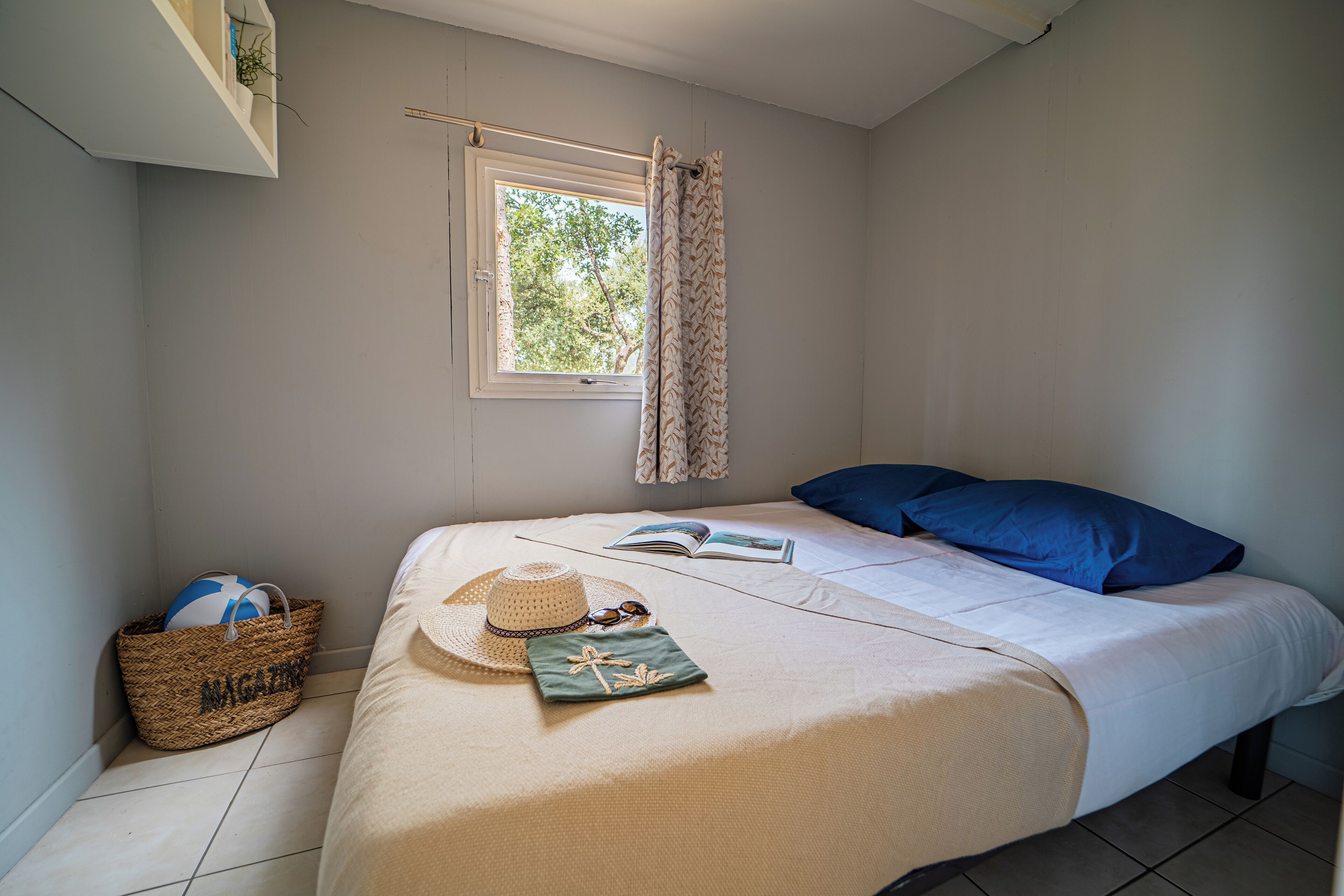 Une chambre avec un lit, une fenêtre avec des rideaux, un panier étiqueté « MAGAZINES », un chapeau, des lunettes de soleil et un livre sur le lit.