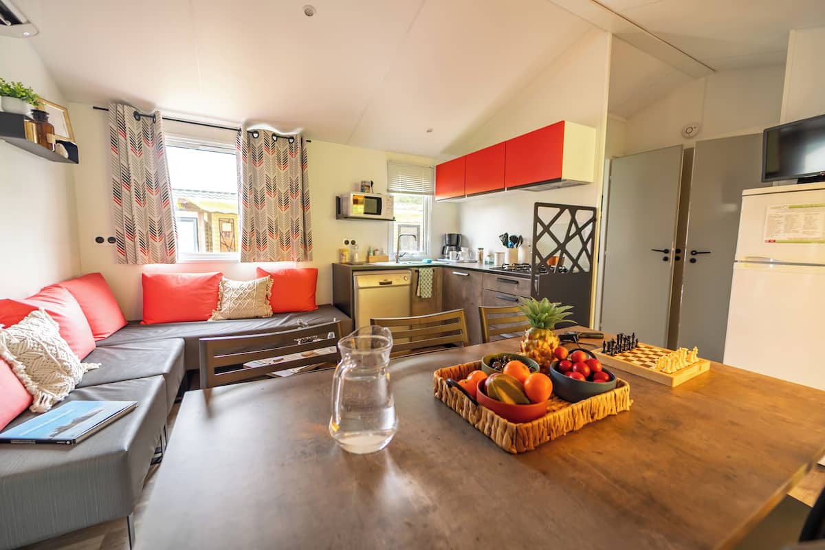 Un espacio habitable pequeño con sofá, una mesa de comedor con fruta y ajedrez, zona de cocina y televisión.