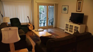 Een woonkamer met een bank, een bijzettafeltje, een gitaar, een televisie op een kast en een schuifdeur naar een buitenruimte.