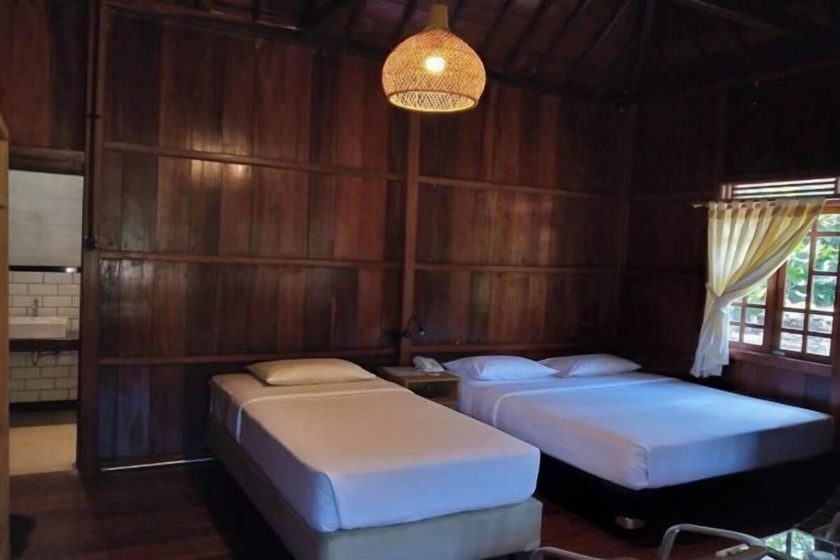 Una habitación con dos camas, paredes de madera, una lámpara colgante, una ventana con cortinas y una mesita de noche pequeña.