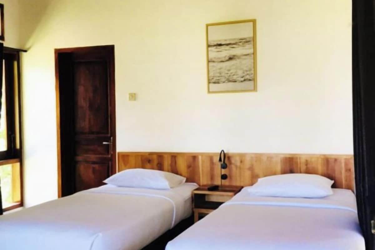 Una habitación con dos camas individuales, cabeceros de madera, una mesita de noche con lámpara y un cuadro enmarcado en la pared.