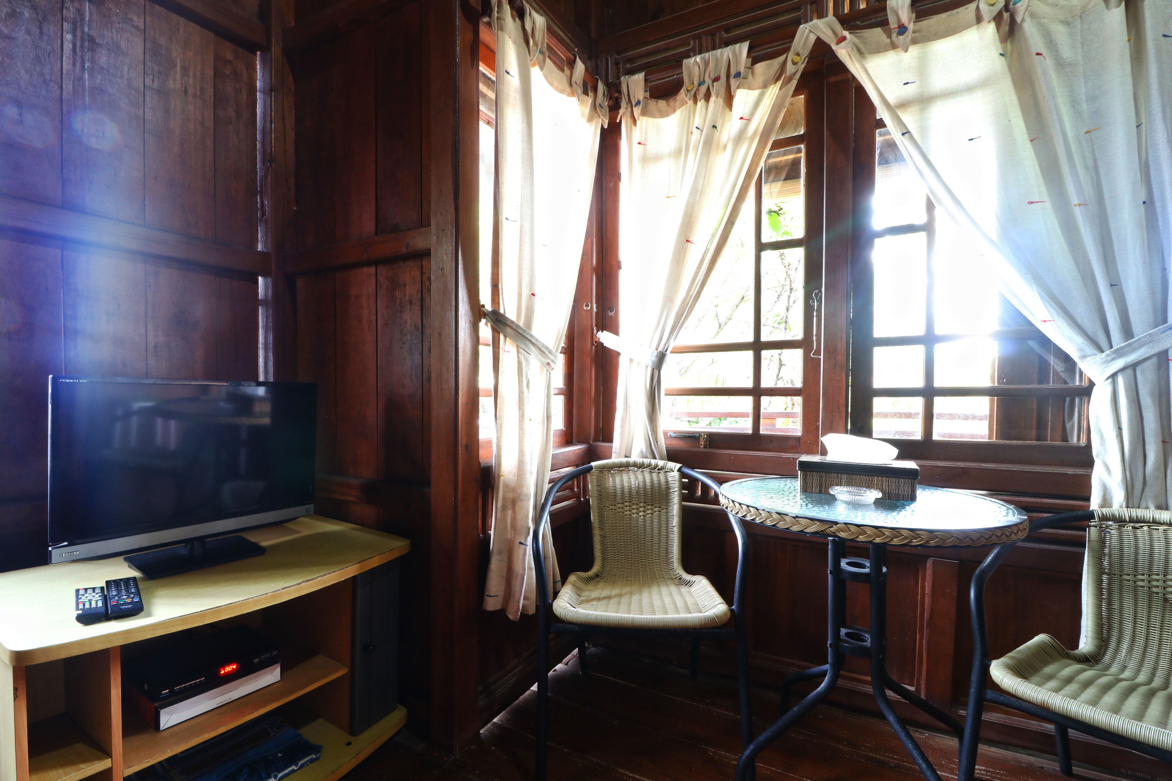 Ein Zimmer mit Holzboden, einem Fernseher auf einem Regal, einem Tisch mit Stühlen und einem Fenster mit Vorhängen.