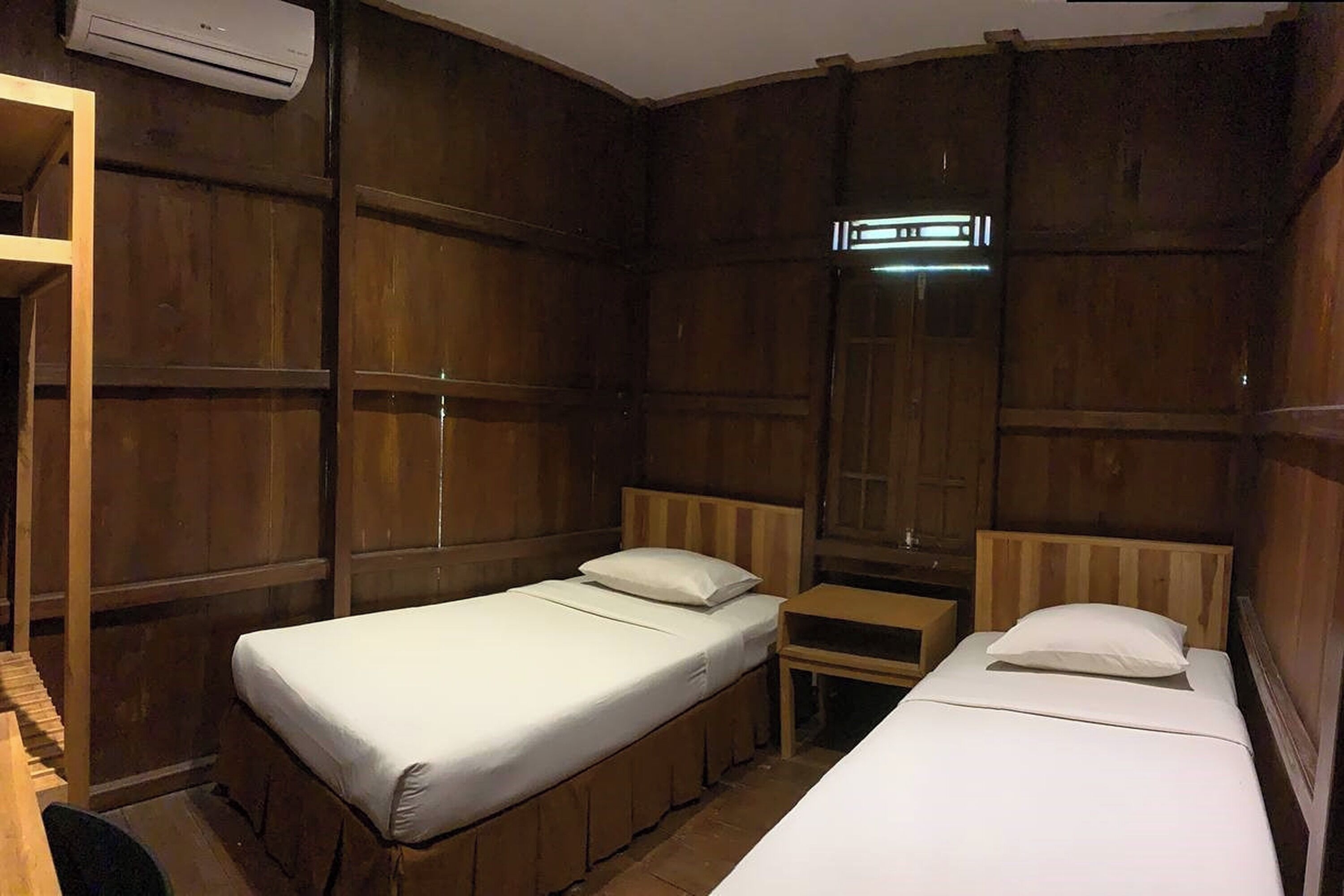 A room with two single beds, wooden paneling on the walls, a small bedside table, and an air conditioning unit.