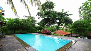 Ein rechteckiger Pool mit Fliesenrand, umgeben von üppiger Vegetation und einigen Gebäuden im Hintergrund.